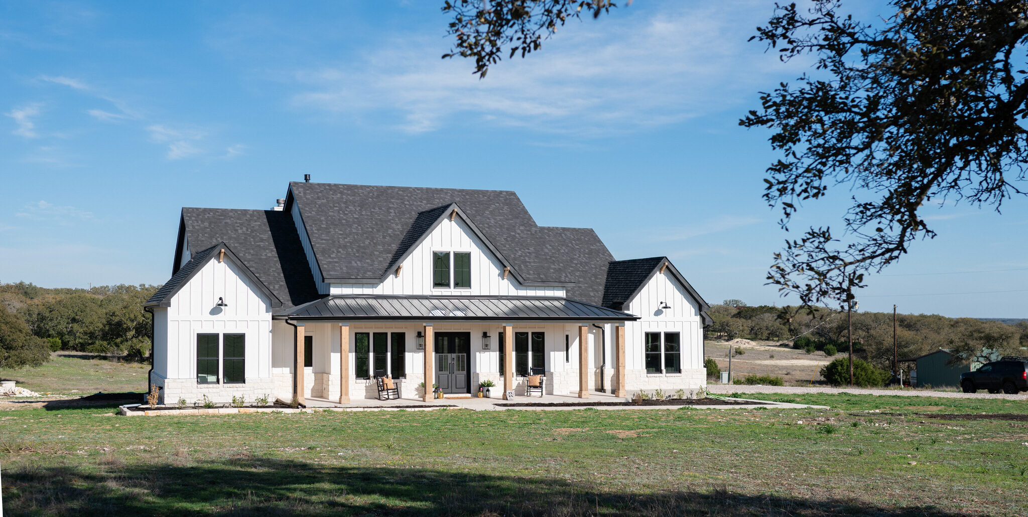 Modern farmhouse-style custom home with white board and batten siding, black roof, and wood accents, built by Jace & Bowen Custom Builders in Burnet, Texas. Surrounded by open Texas Hill Country landscape under a clear blue sky.